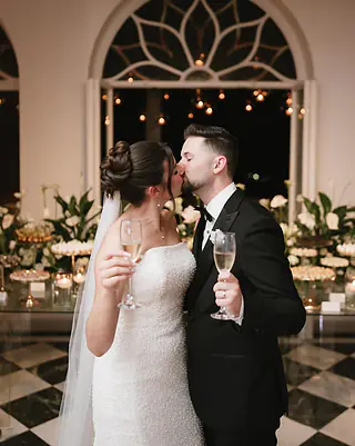 Noivos se beijando e erguendo taças de espumante diante da mesa de doces, piso xadrez e janela em arco