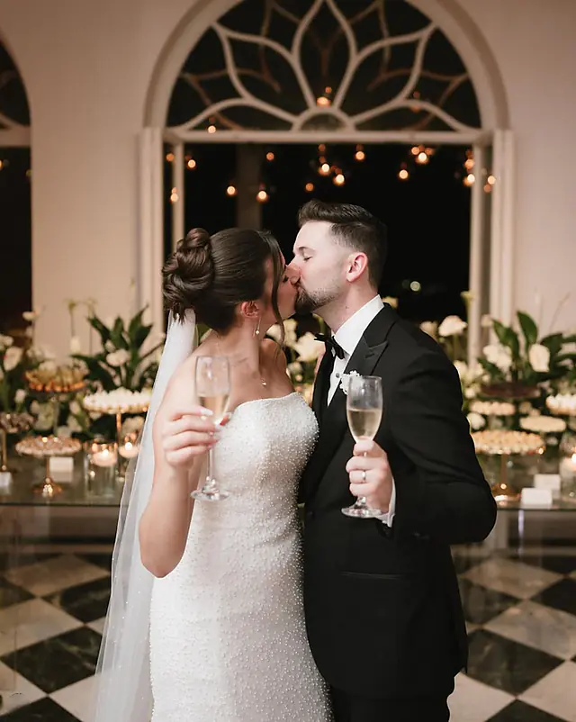 Noivos se beijando e erguendo taças de espumante diante da mesa de doces, piso xadrez e janela em arco