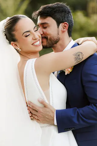 Noivo abraça e beija o rosto da noiva, que sorri de olhos fechados