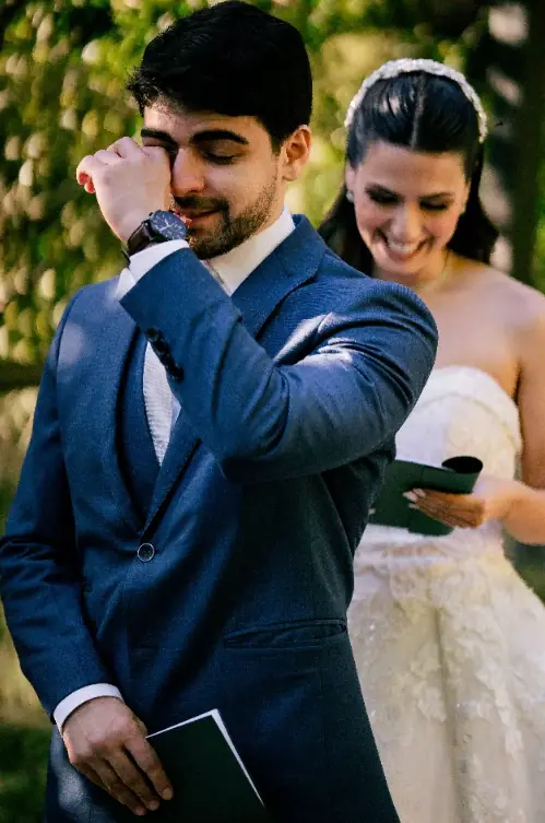 Noivo enxuga as lágrimas segurando uma carta, com a noiva sorrindo logo atrás